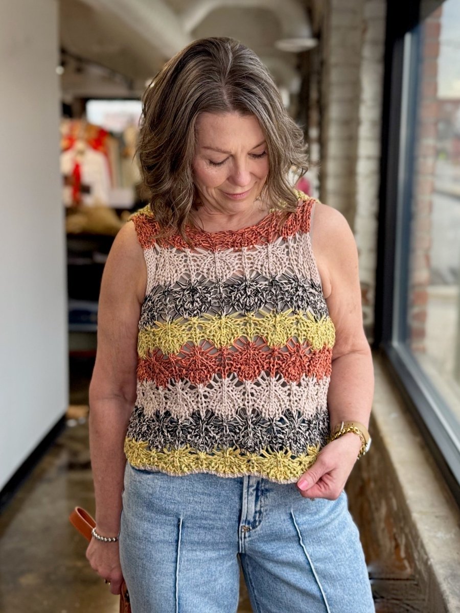 A woman wearing a sleeveless, striped crochet knit top with horizontal bands of rust orange, gray, yellow, and cream, paired with light-wash blue jeans, standing in a shop interior.