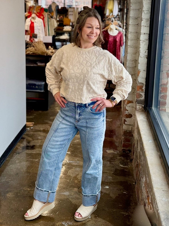 A woman modeling a cream cable-knit sweater and light-wash blue jeans in a retail store setting.