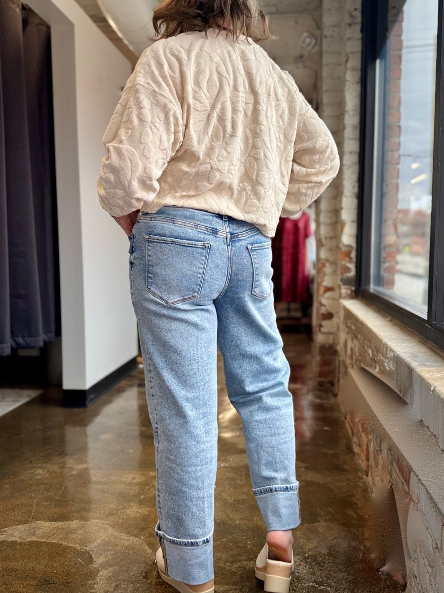 A person shown from behind wearing a cream textured cropped jacket and light blue cuffed jeans in a retail store hallway.