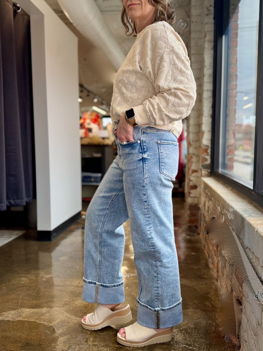 A woman wearing light-wash wide-leg denim jeans with rolled cuffs, paired with a cream textured long-sleeve top and beige platform slide sandals, standing in a retail store interior.