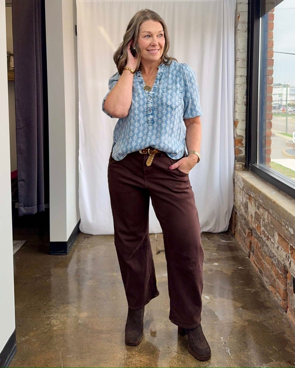 Woman wearing a blue patterned blouse and brown pants standing indoors.