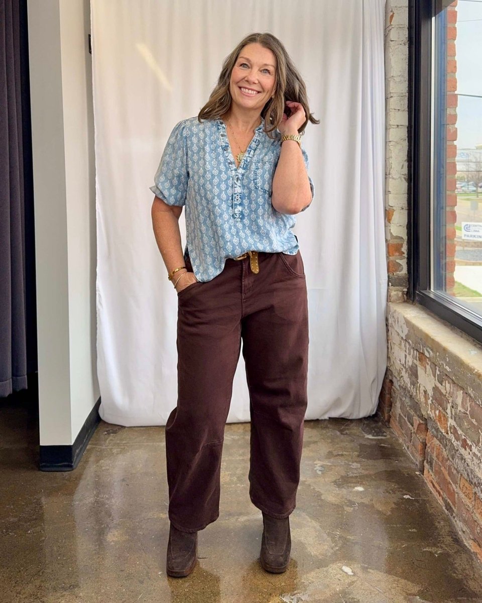 Woman wearing a light blue patterned shirt and brown pants standing indoors.