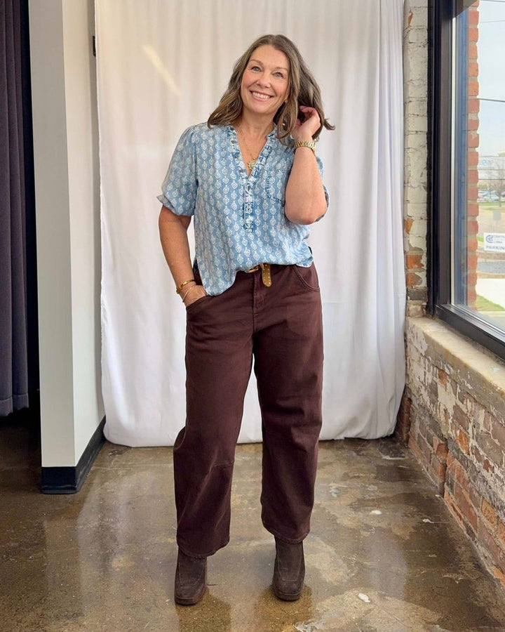 Woman wearing a light blue patterned shirt and brown pants standing indoors.