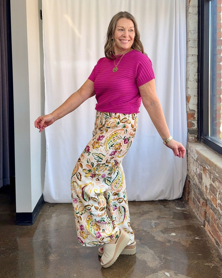 Woman wearing a bright pink top and colorful patterned pants standing indoors.