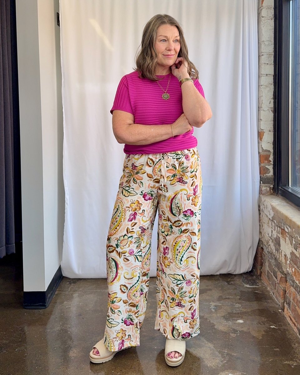 Woman wearing a pink top and floral pants standing against a white curtain.