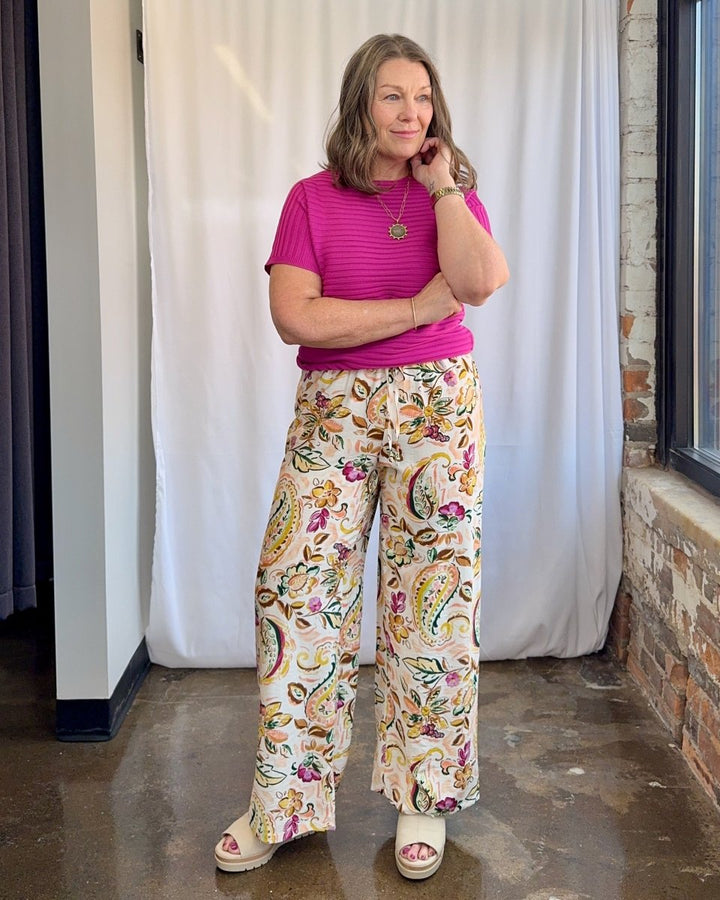 Woman wearing a pink top and floral pants standing against a white curtain.