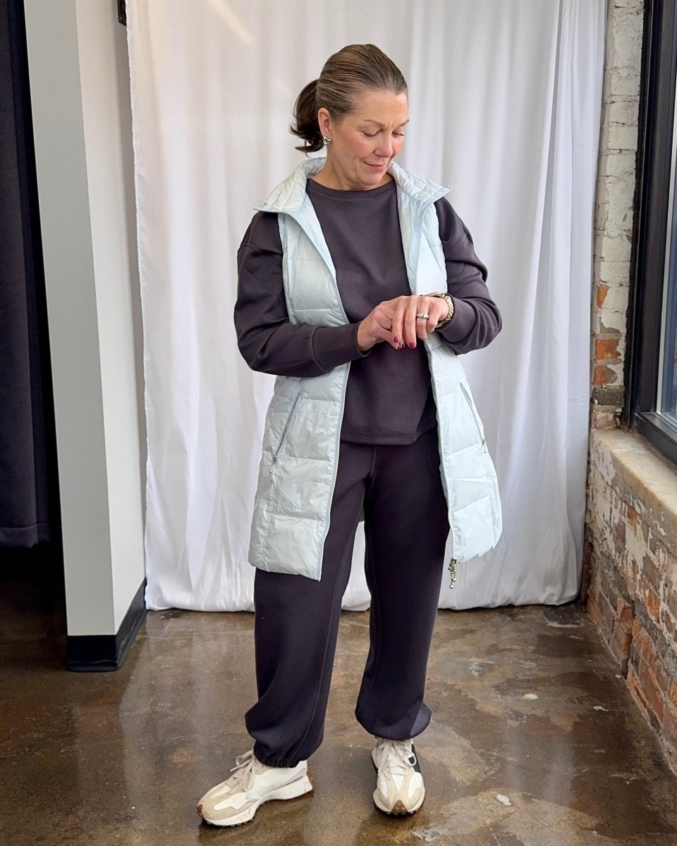 Woman wearing a gray outfit with a white puffer vest against a white curtain background.