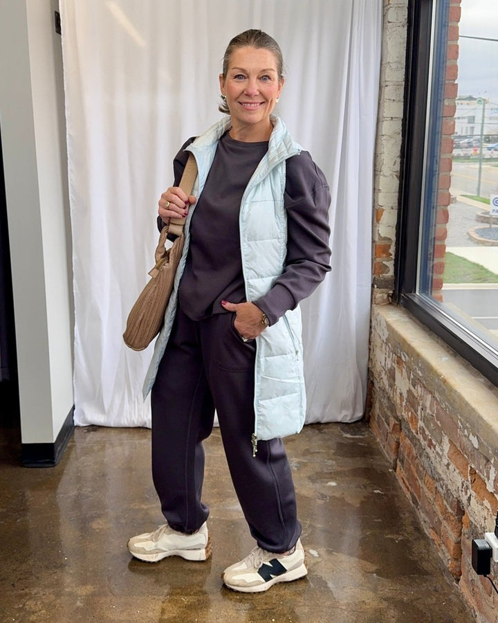 Person wearing a white puffer vest over a dark outfit, standing indoors with a window and brick wall in the background.