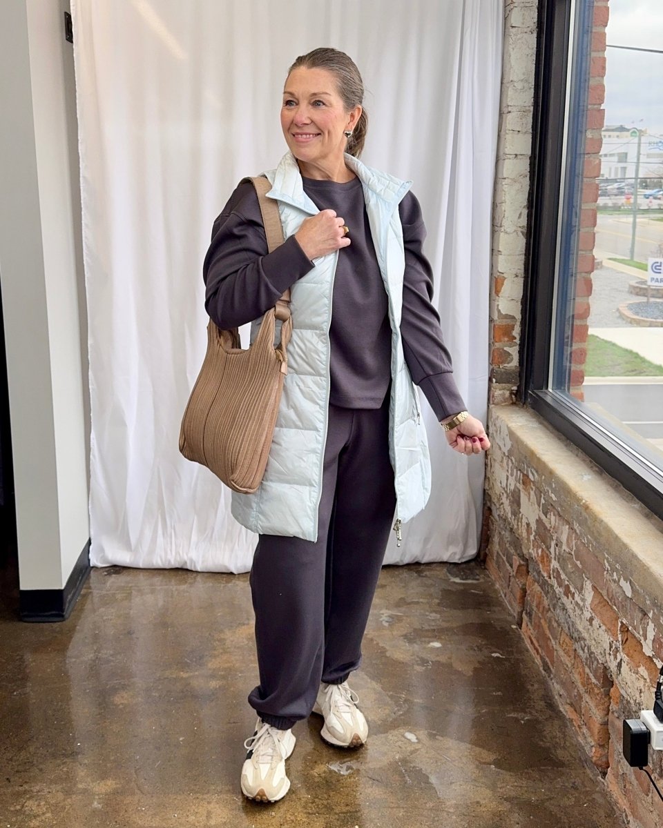Woman wearing a white puffer vest over a dark outfit, holding a beige bag, standing indoors.