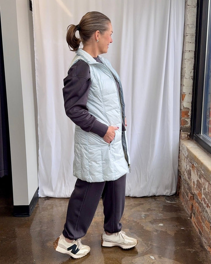 Person wearing a light blue puffer vest over a dark jacket, standing against a white curtain.