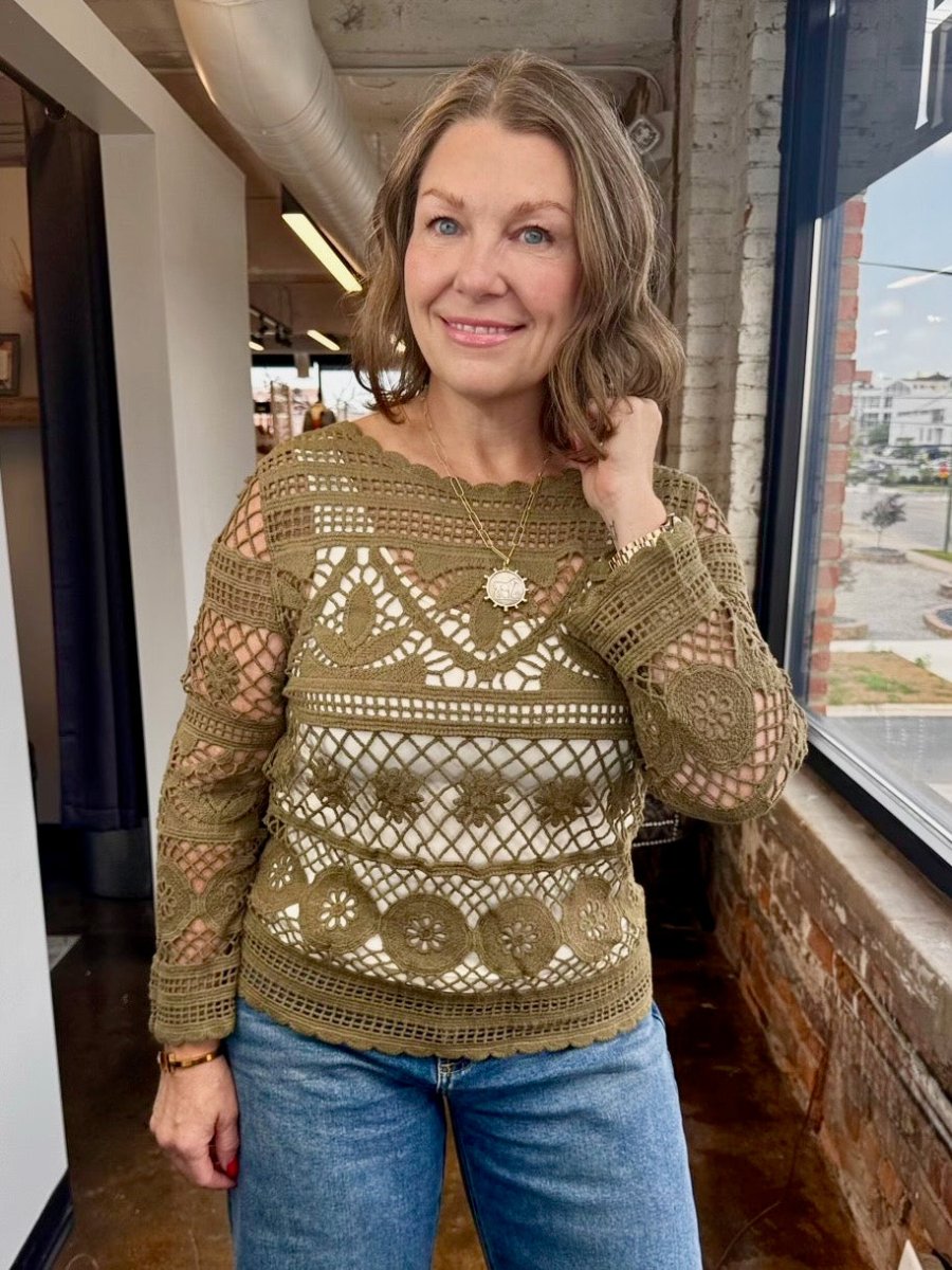 A woman wearing a brown crochet knit long-sleeve top with blue jeans, standing in an indoor loft space with brick walls and a window.