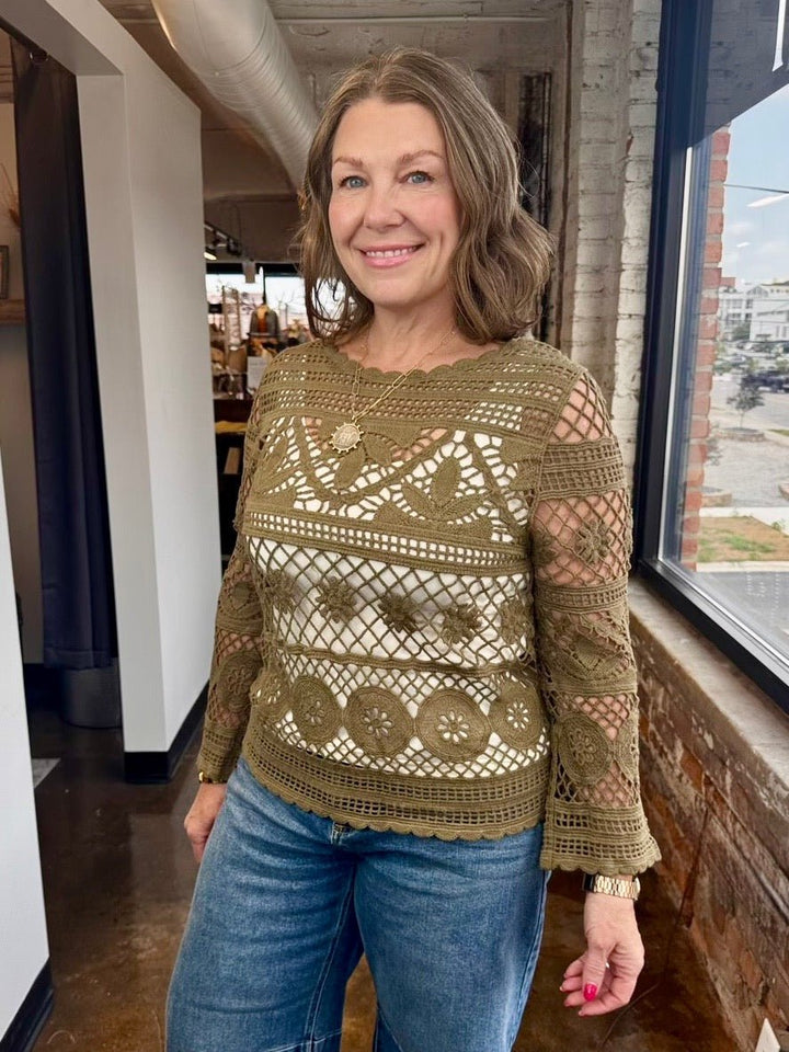 A woman wearing a brown crochet knit long-sleeve top with blue jeans, standing in a modern indoor space with a large window.