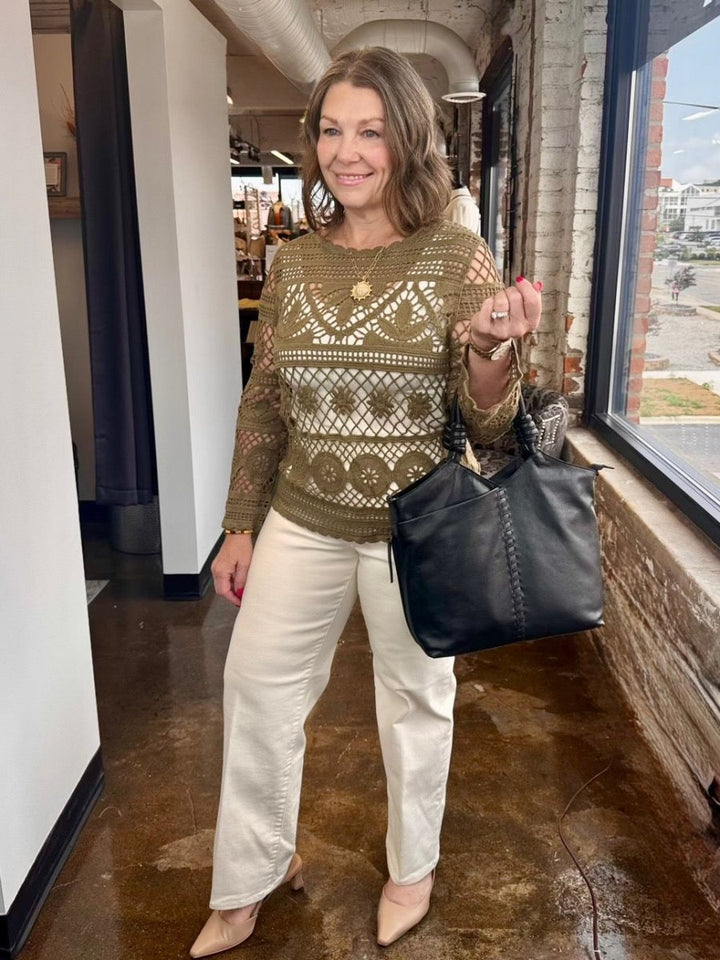 A woman stands in a shop corridor wearing a textured olive green and brown knit sweater with a geometric pattern, paired with cream wide-leg pants and beige heels, while holding a large black leather tote bag.