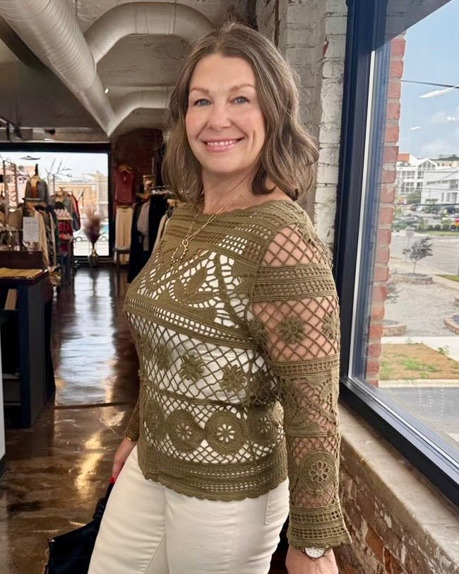 A woman wearing a brown crochet knit top with white pants, standing in a shop next to a window.