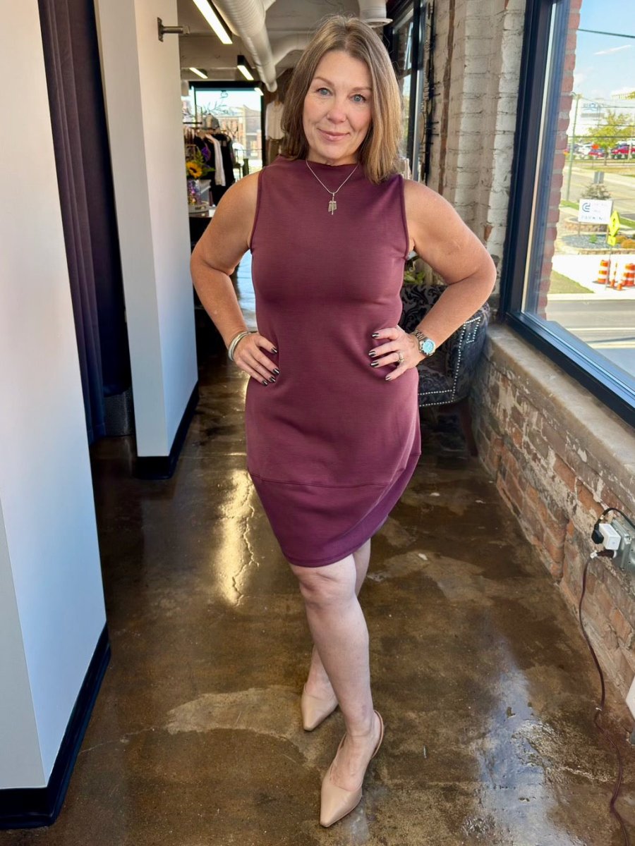 A woman modeling the Juno Dress by Revir in a rich plum berry shade, featuring a sleeveless high-neck bodice and a flowing midi skirt for a polished, effortless look.