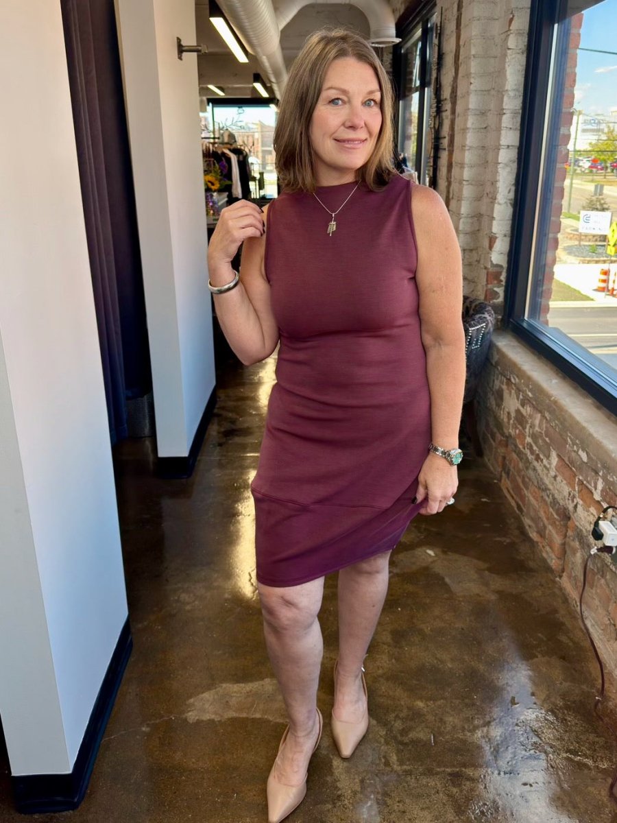 A woman modeling the Juno Dress by Revir in a rich plum berry shade, featuring a sleeveless high-neck bodice and a flowing midi skirt for a polished, effortless look.