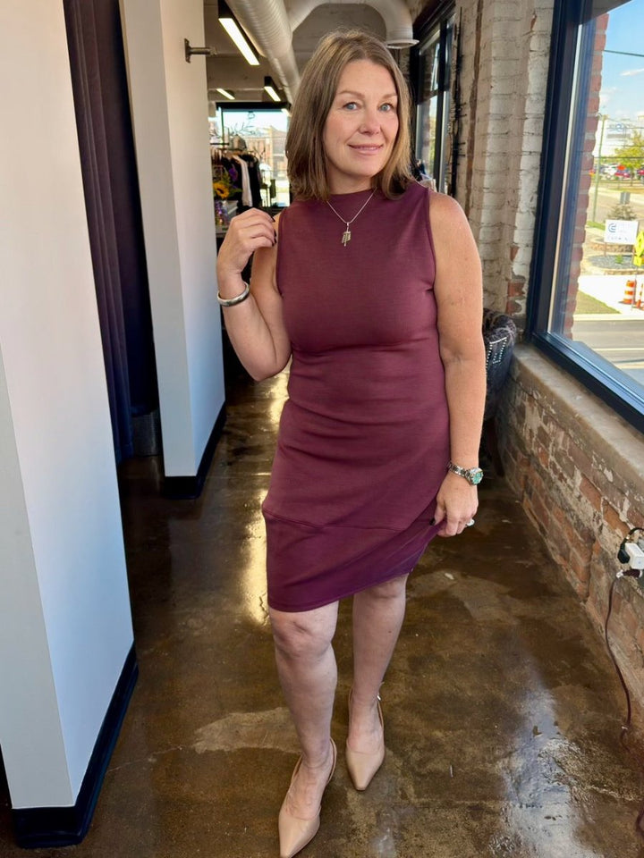 A woman modeling the Juno Dress by Revir in a rich plum berry shade, featuring a sleeveless high-neck bodice and a flowing midi skirt for a polished, effortless look.