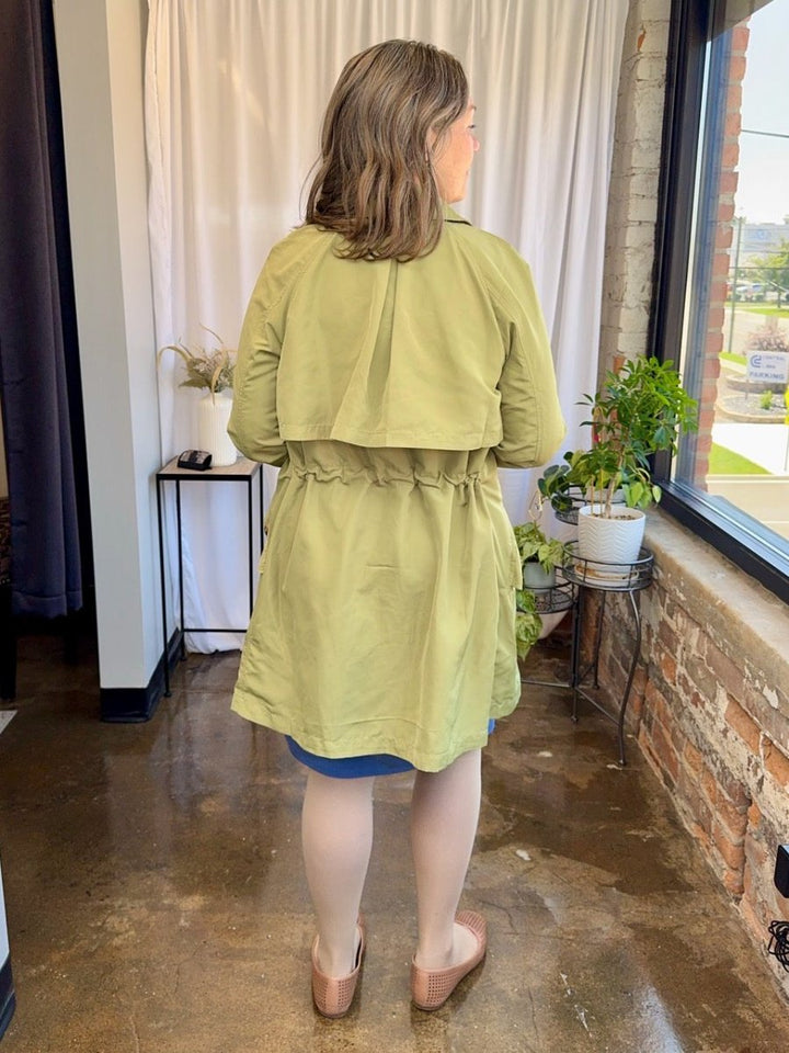 Person wearing a light green dress standing in a room with plants and a window.