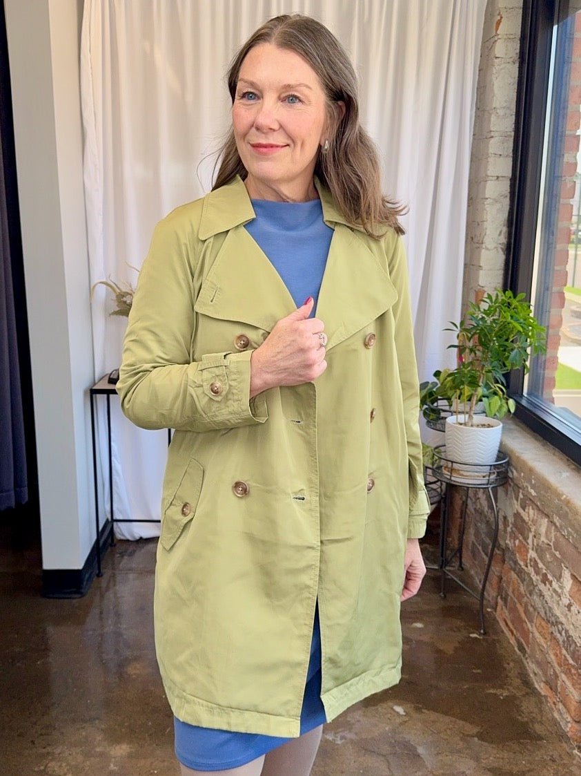 Woman wearing a green trench coat indoors with a plant and window in the background