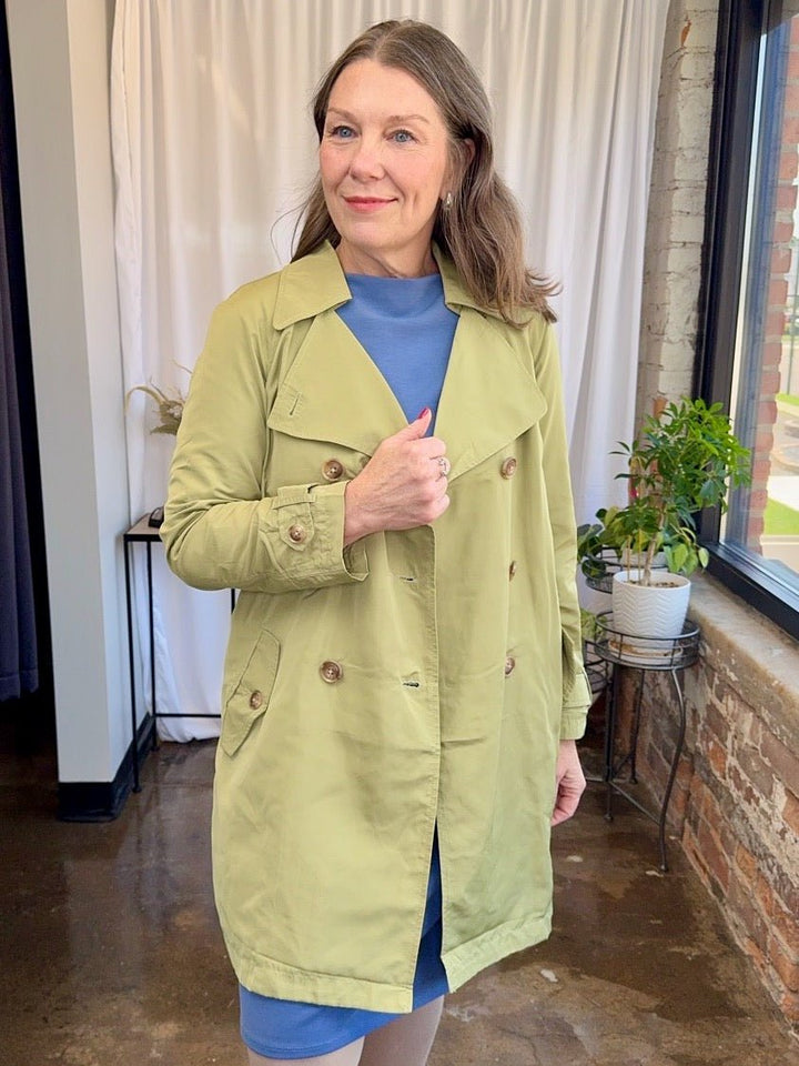 Woman wearing a green trench coat indoors with a plant and window in the background