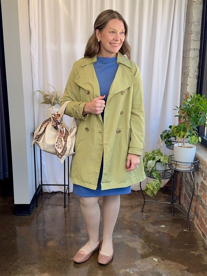 Woman in a green trench coat standing indoors with plants and a curtain in the background