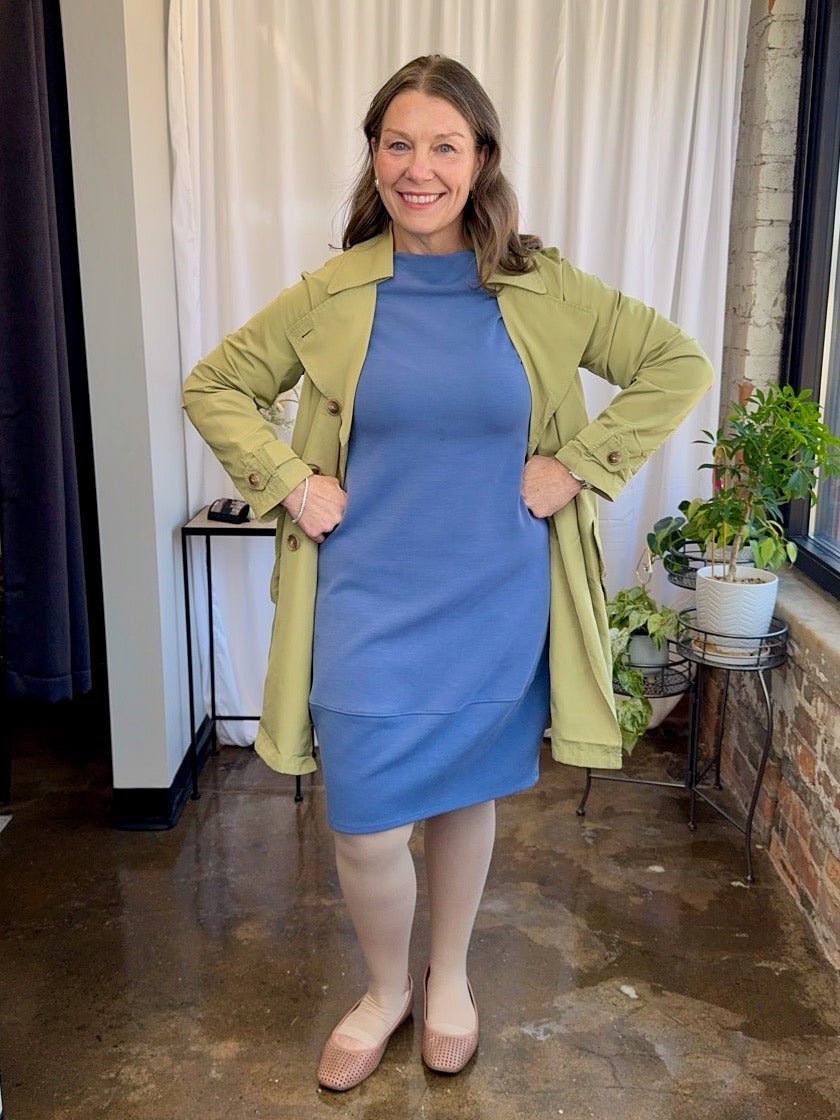Woman wearing a blue dress and green coat indoors with plants and furniture in the background.
