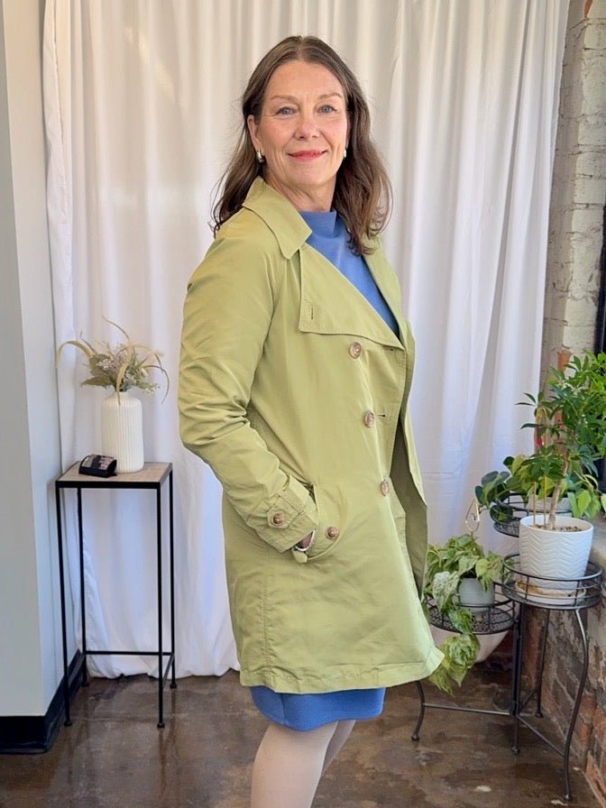 Woman wearing a green jacket standing in a room with plants and a white curtain.