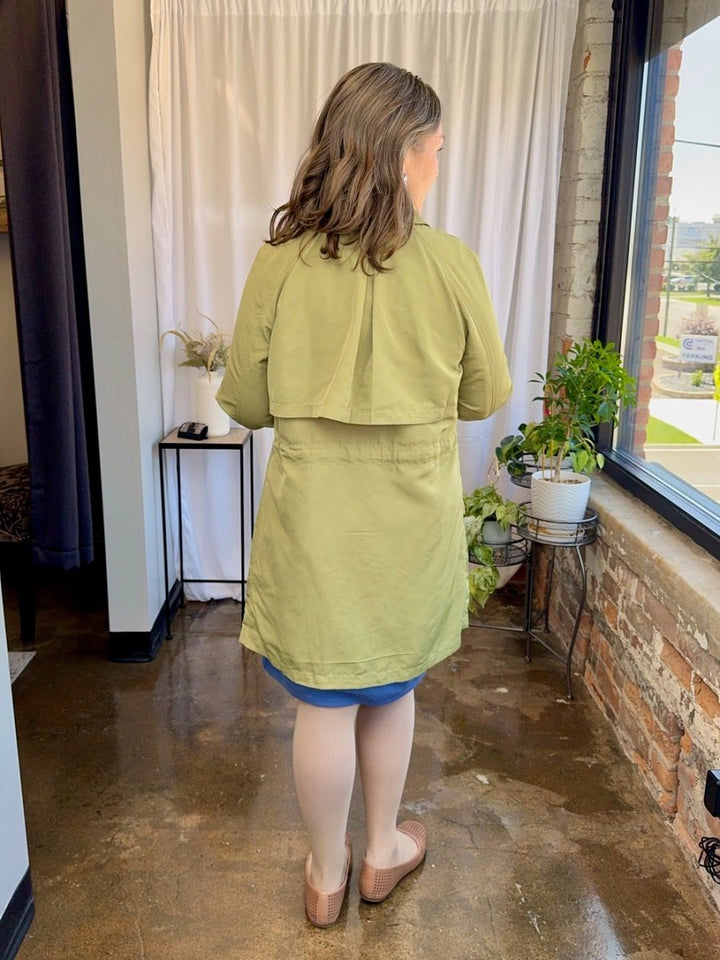 Woman wearing a green jacket standing in a room with a window and plants.