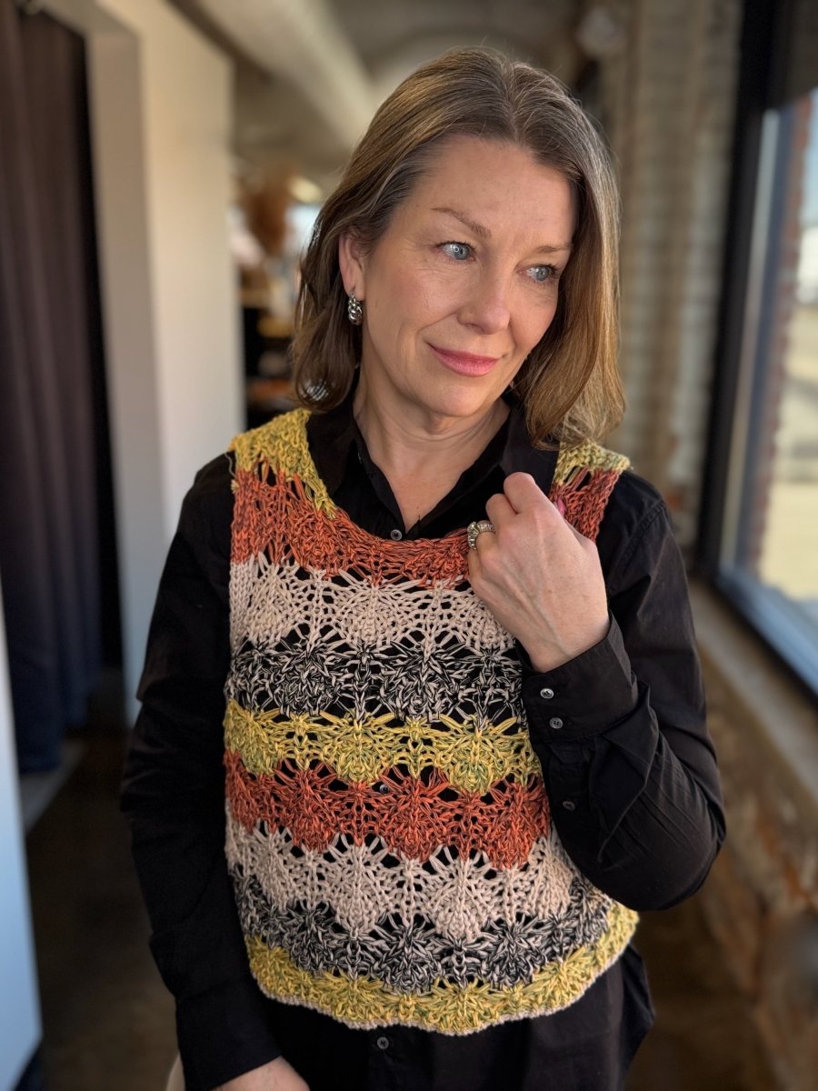 A woman wearing a black long-sleeve button-up shirt and a colorful crocheted vest with horizontal stripes of yellow, orange, black, and cream.