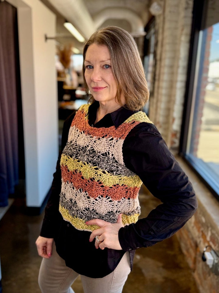 A woman wearing a colorful striped crochet vest over a black long-sleeve shirt and beige pants, standing in a bright indoor hallway with brick walls and large windows.