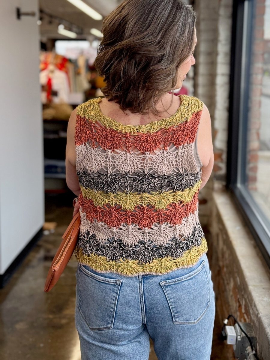 A woman viewed from behind wearing a sleeveless, striped crochet knit top with horizontal bands of mustard yellow, rust red, cream, and gray, paired with light blue jeans.
