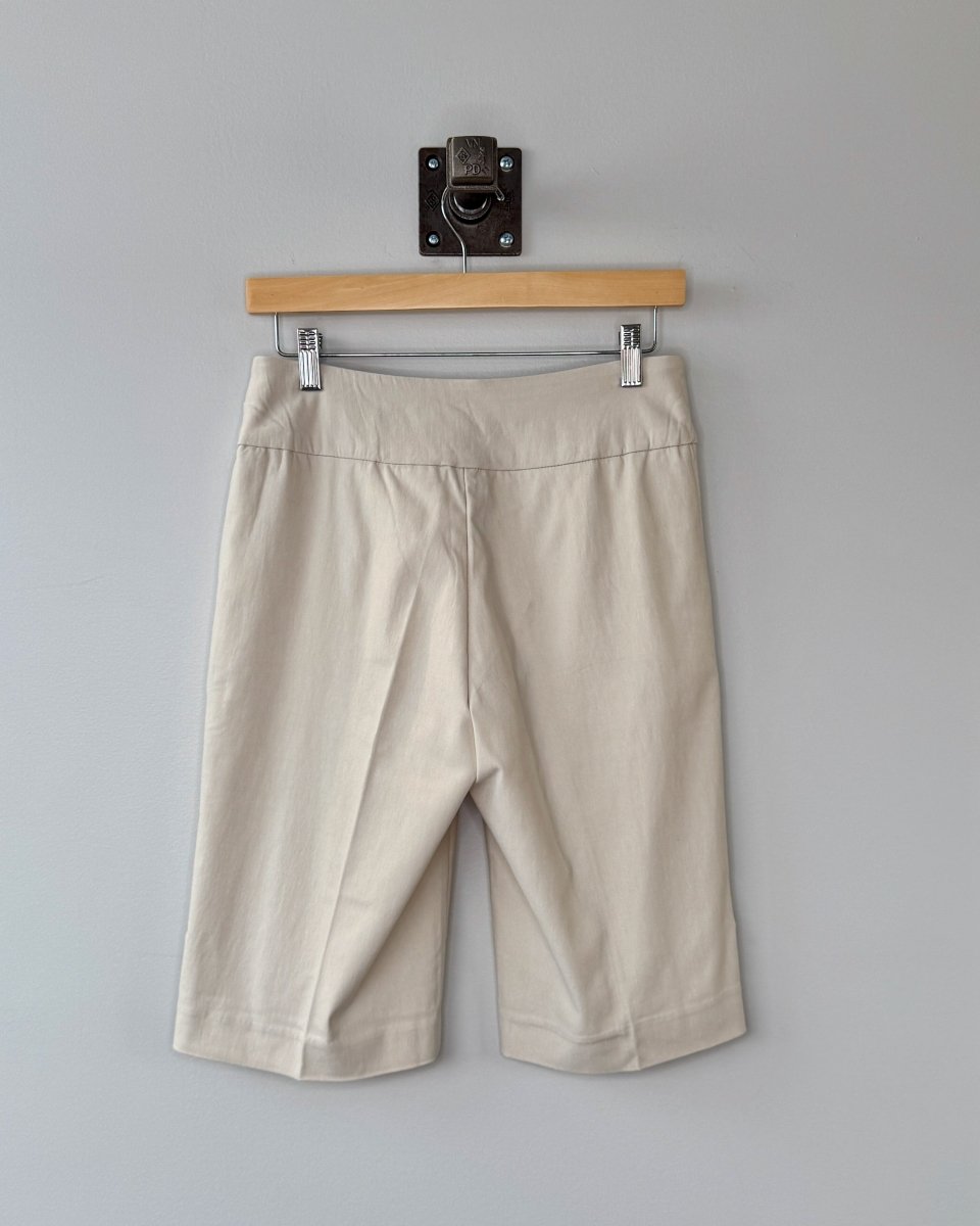 Beige shorts hanging on a wooden hanger against a gray wall.