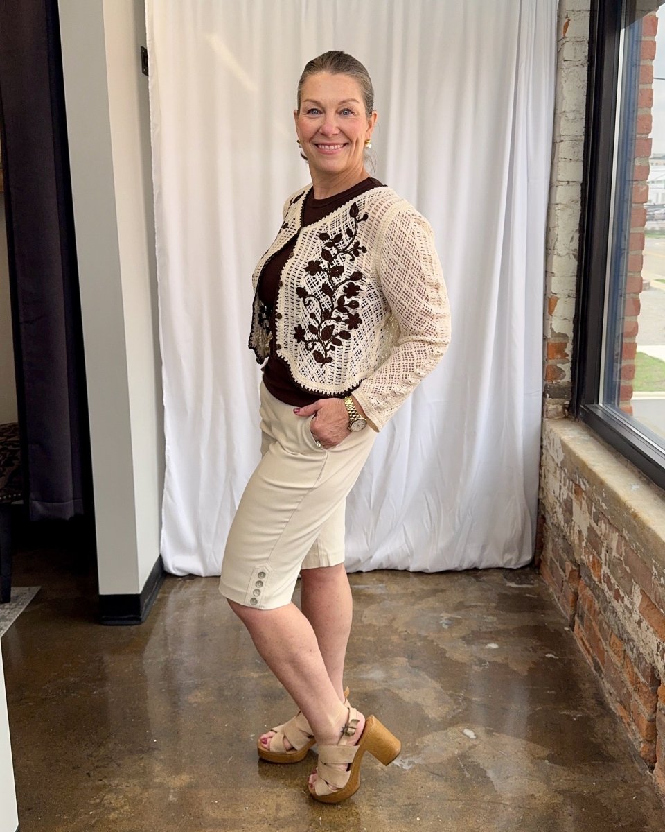 Woman wearing a patterned sweater and shorts standing indoors with a white curtain and brick wall in the background.