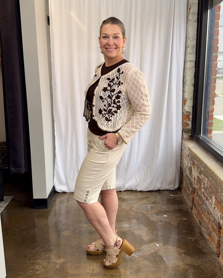 Woman wearing a patterned sweater and shorts standing indoors with a white curtain and brick wall in the background.