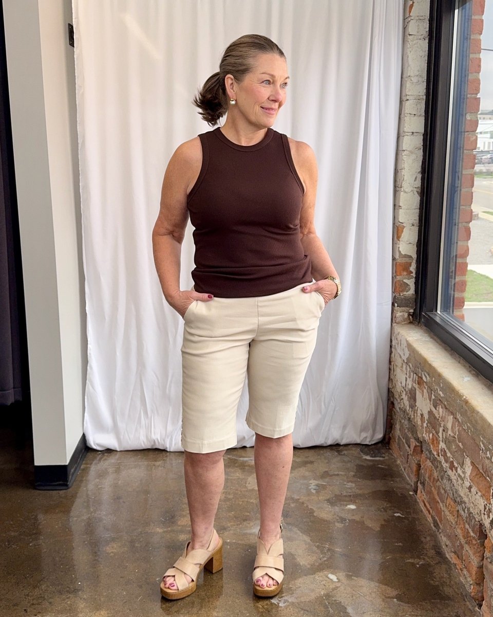 Woman wearing a brown tank top and shorts standing indoors with a white curtain and brick wall in the background.
