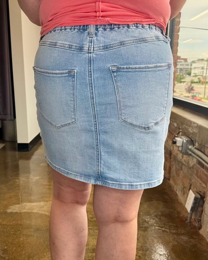 A person shown from the back wearing a light-wash denim mini skirt with two large patch pockets and a coral pink top, standing indoors near a window.