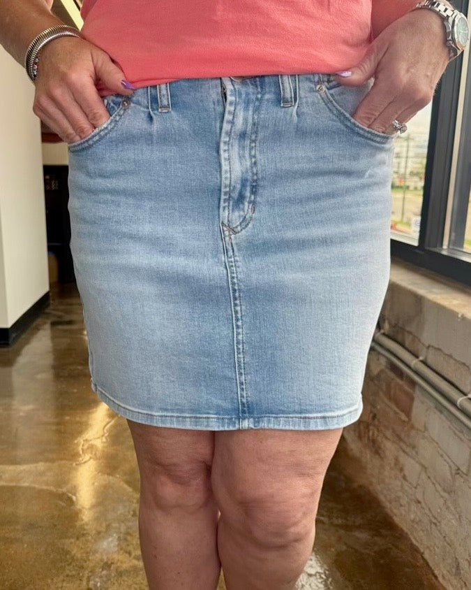A person wearing a light-wash blue denim mini skirt with a pink top, standing indoors near a window.