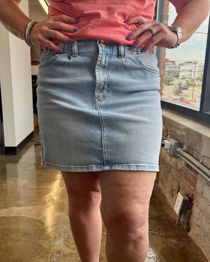 A person wearing a light-wash blue denim mini skirt with a peach-pink top, standing indoors near a window.