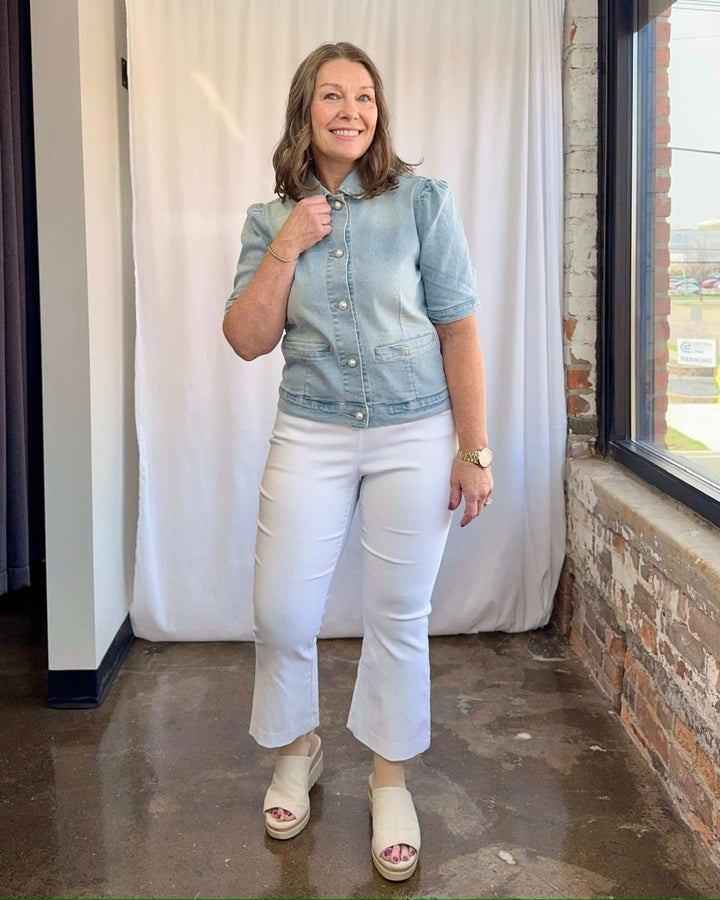 Woman wearing a denim jacket and white pants standing in front of a white curtain.