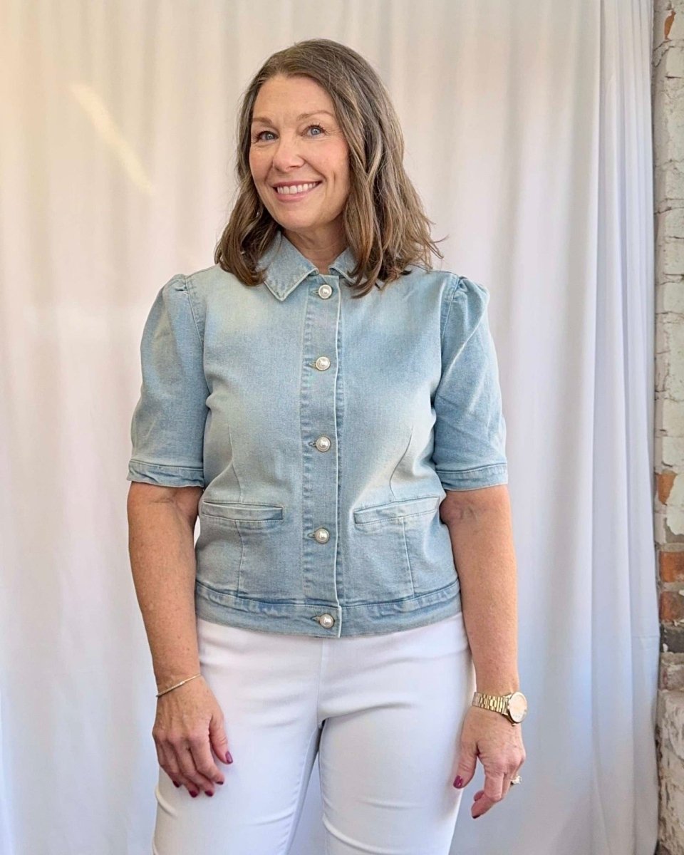 Woman wearing a light blue denim jacket and white pants standing against a white curtain.