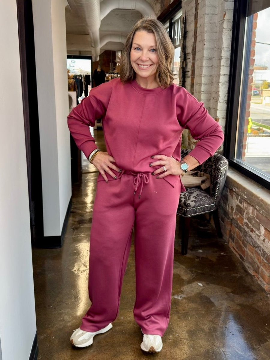A woman wearing a matching dusty rose pink lounge set consisting of a long-sleeve sweatshirt and drawstring pants, standing in a storefront hallway.