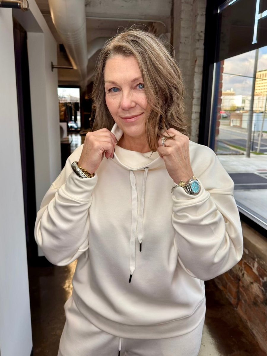 A woman wearing a cream-colored matching sweatshirt and sweatpants set, posing in front of a window in an industrial loft setting.