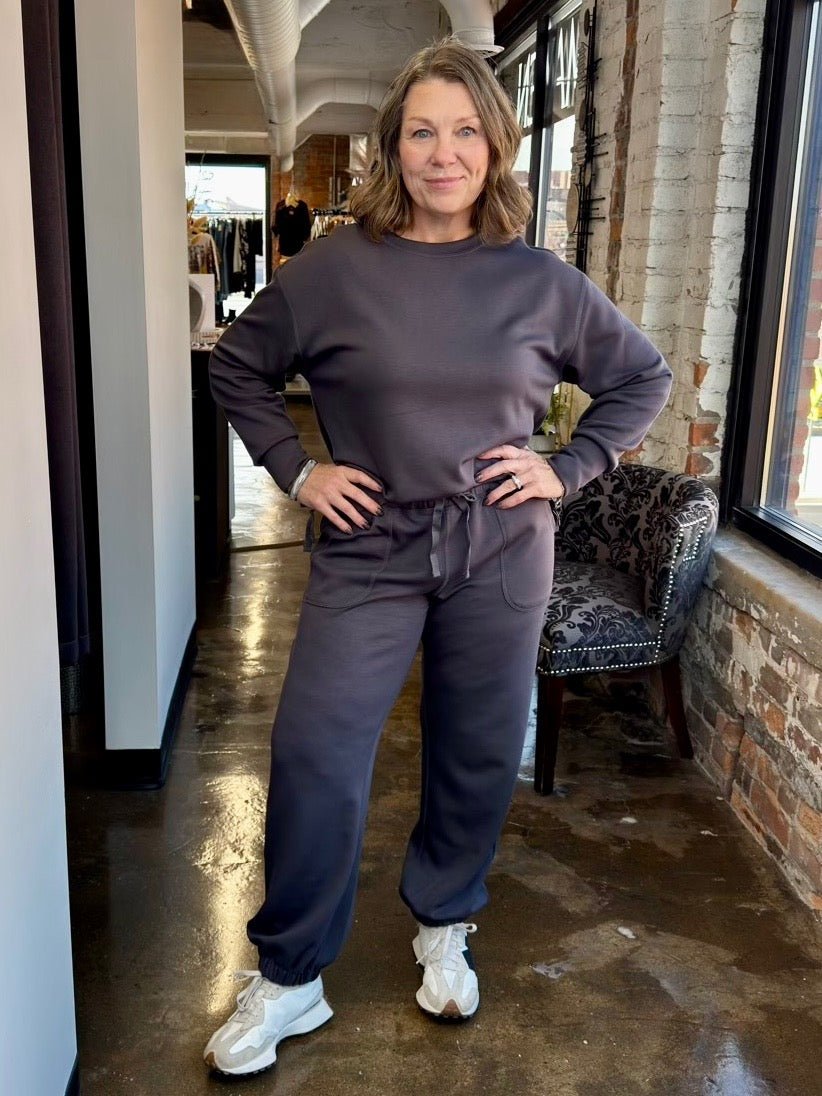 A woman modeling a matching dark gray sweatshirt and jogger pants set with white sneakers, standing in a shop interior with brick walls and windows.