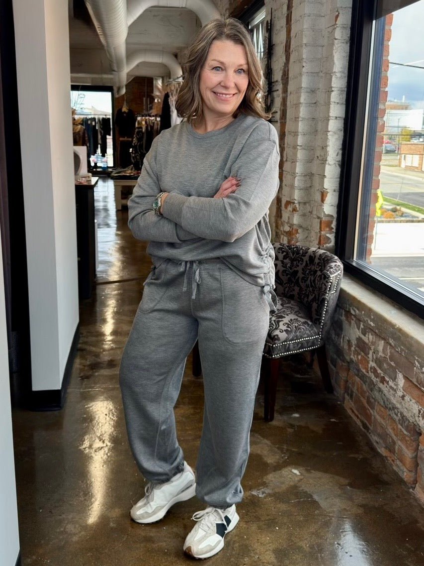 A woman wearing a matching gray sweatshirt and jogger pants set, standing in a brick-walled boutique hallway with her arms crossed.