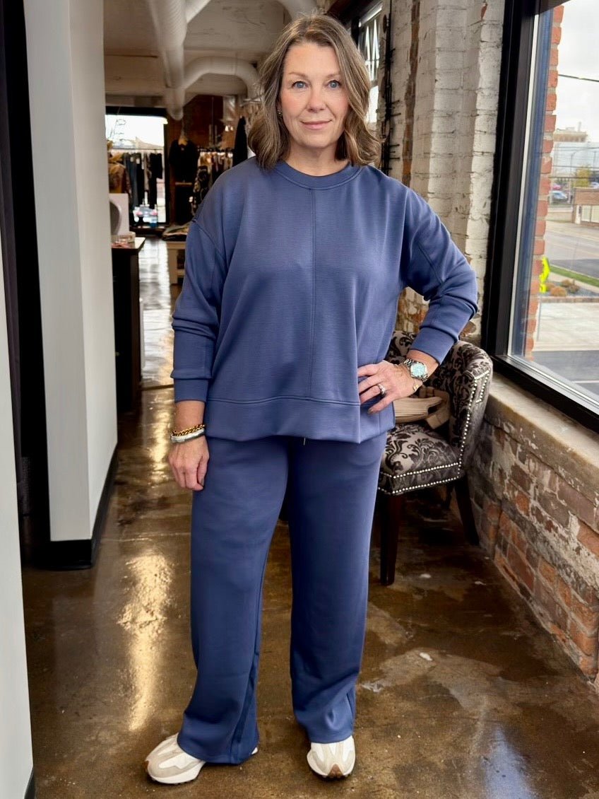 A woman modeling a matching navy blue sweatshirt and sweatpants lounge set in a retail store setting with a brick wall and large window.