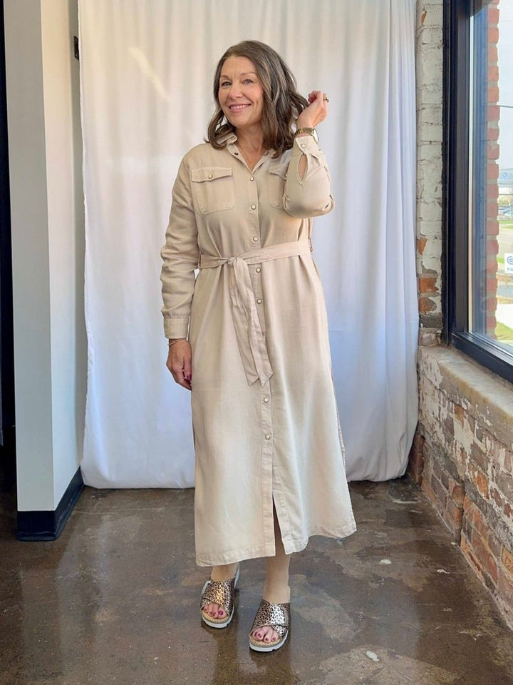 Woman wearing a beige dress standing indoors with a white curtain and brick wall in the background.
