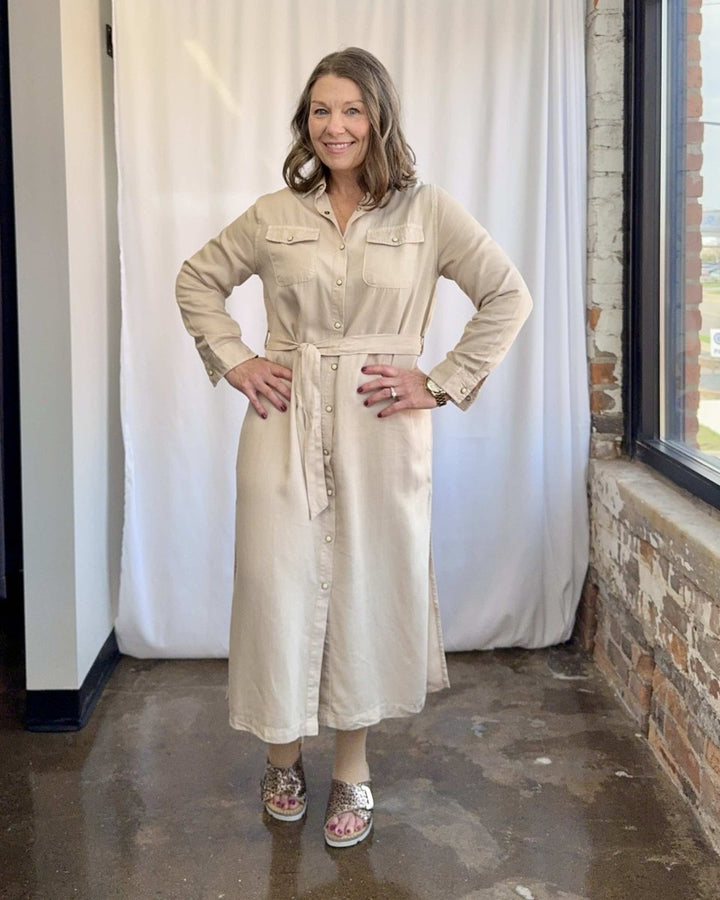 Woman wearing a beige dress standing in a room with white curtains and a brick wall.