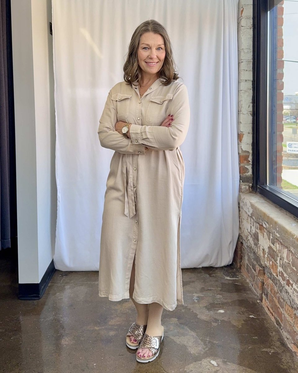 Woman wearing a beige trench coat standing indoors with a white curtain and brick wall in the background.