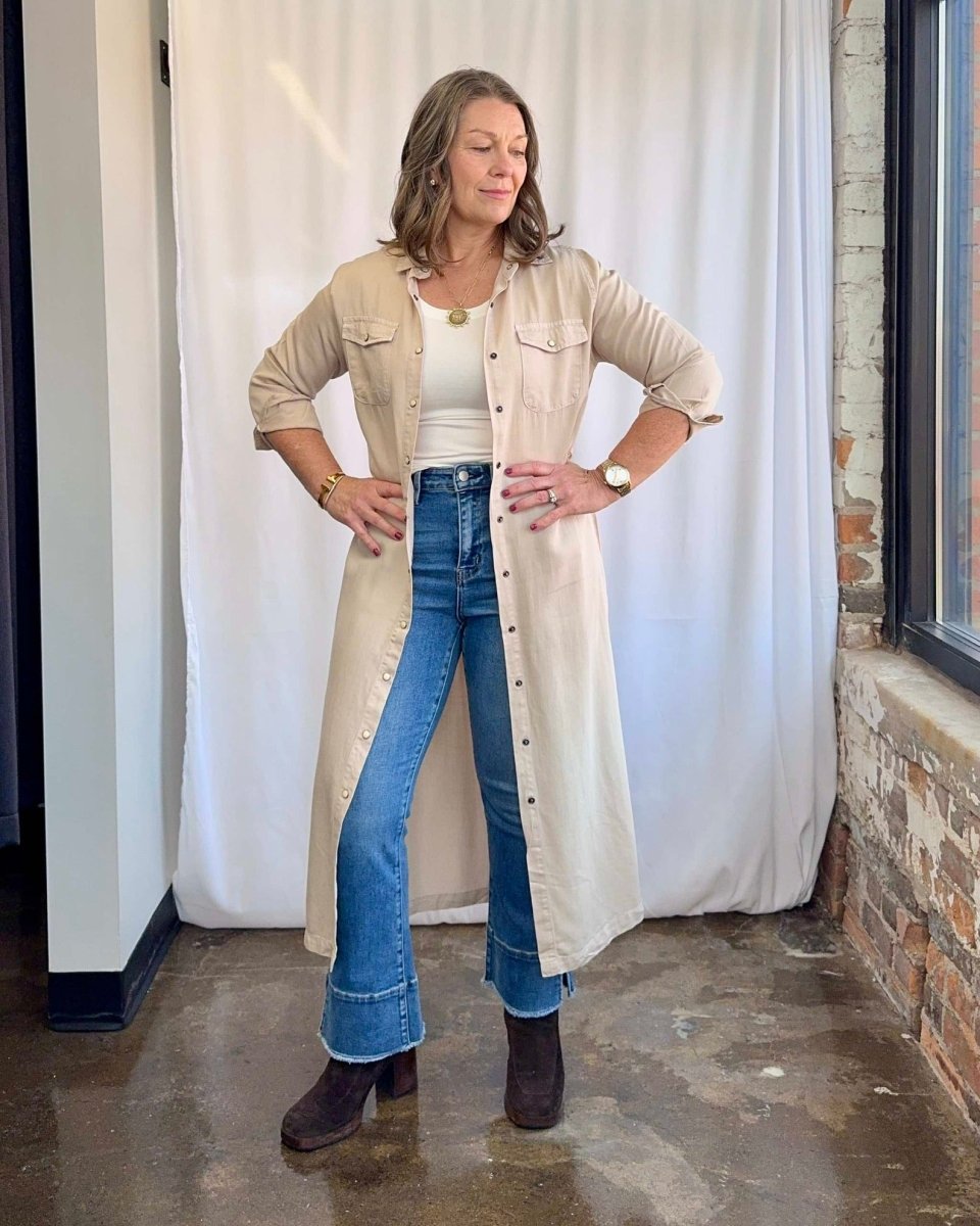 Woman wearing a beige long coat, white top, blue jeans, and brown shoes indoors.