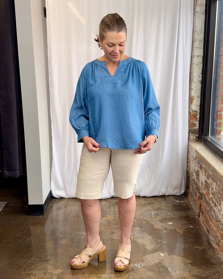 Woman wearing a blue blouse and beige shorts standing indoors with a white curtain and brick wall in the background.